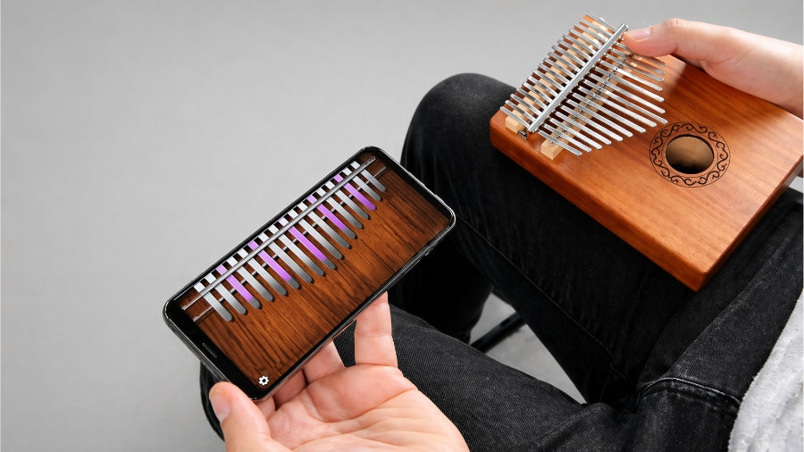 Man holding Android kalimba app on smartphone and real kalimba instrument with light grey studio background