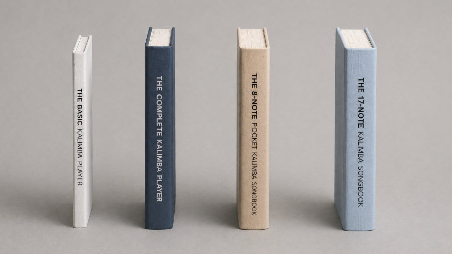 Four kalimba books of varying thickness arranged upright on a soft grey background, showing a thin beginner guide, a medium complete guide, and two thicker songbooks with textured covers and clean vertical titles.
