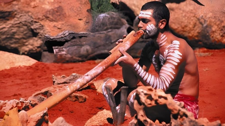 A man playing a didgeridoo, a traditional Indigenous Australian wind instrument, holding it horizontally while blowing into one end.