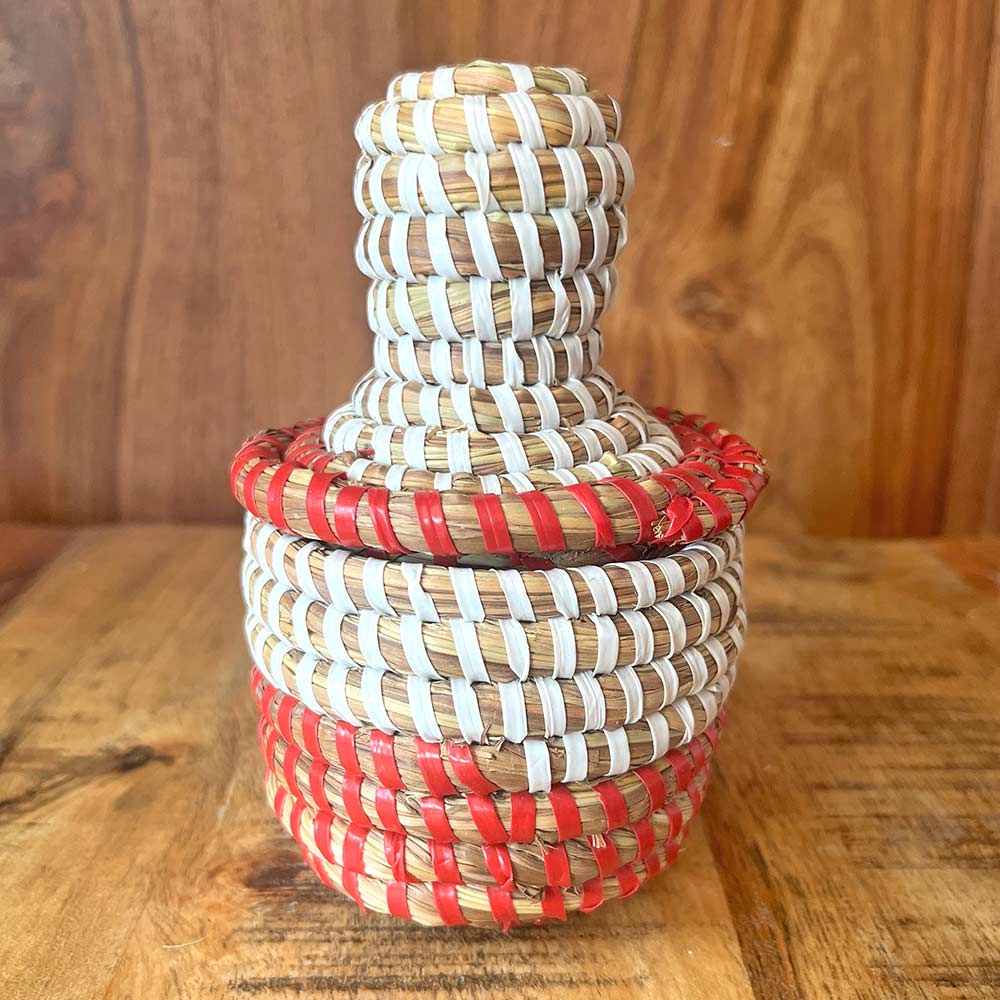 A small, handwoven African basket in red and white, made from straw and recycled plastic, with a matching lid.