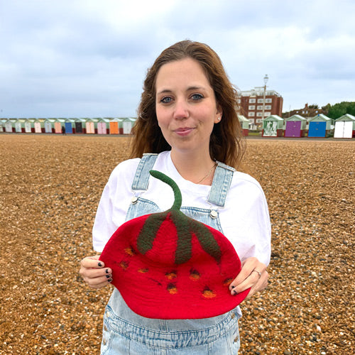Woman holding a red felt strawberry hat on a beach with colorful buildings in the background