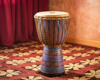 African djembe drum standing upright on a colorful floral carpet, featuring a carved wooden body and a taut natural skin drumhead.