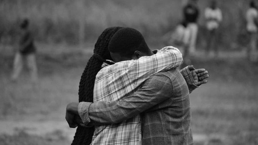 Black and white image of a couple in love hugging, their faces hidden, with people in the background looking on.