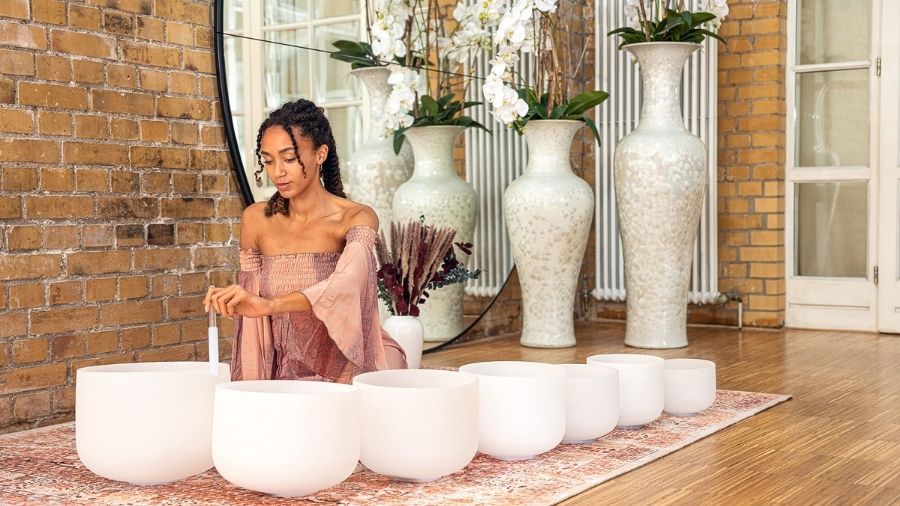 Girl with seven crystal singing bowls of different sizes, holding a mallet and playing one of the bowls.