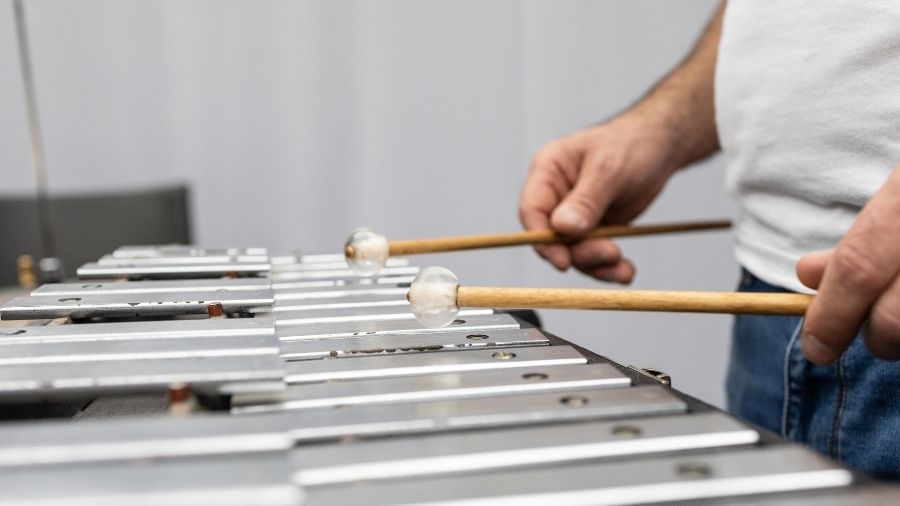 Person playing a glockenspiel with mallets, wearing a white shirt and jeans, focusing on the hands and instrument.