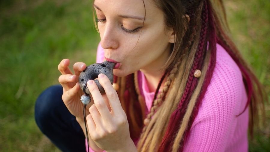 Portrait of a young female shaman in a pink knitted sweater playing a ceramic ocarina in a forest during autumn or spring, creating a relaxing and tranquil scene.