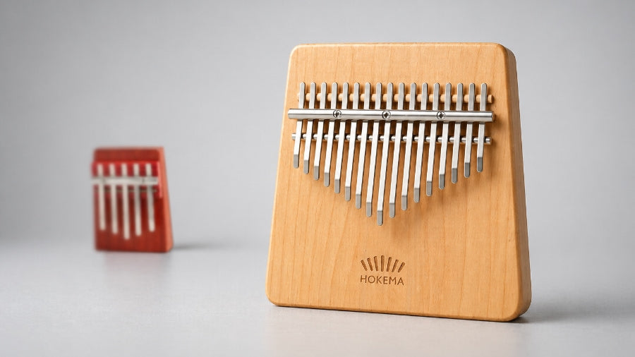 Light wood 17-note premium kalimba in sharp focus with a blurred red 5-note beginner kalimba behind on a silver grey studio background.