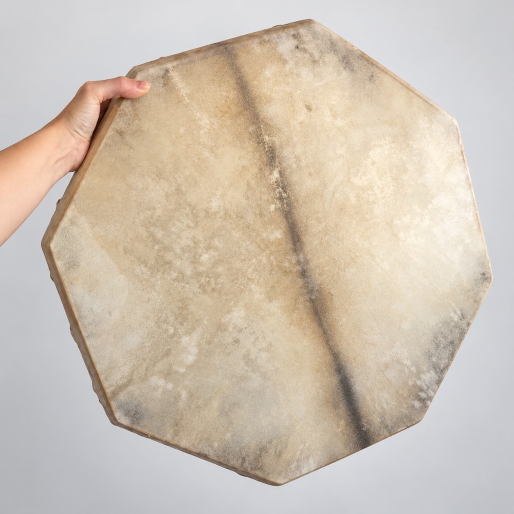 A hand holds a large octagonal drum with a natural beige and brown animal skin head against a plain light gray background.
