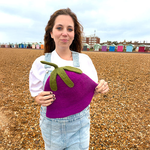 Woman holding a purple eggplant-shaped hat on a beach with colorful huts in the background