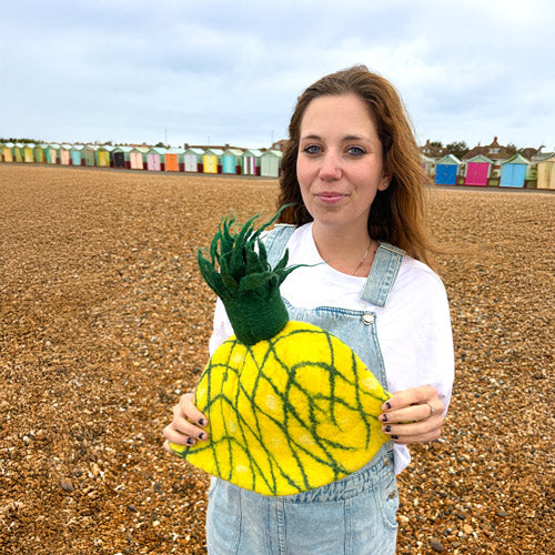 Woman holding a pineapple-shaped hat on a beach with colorful huts in the background