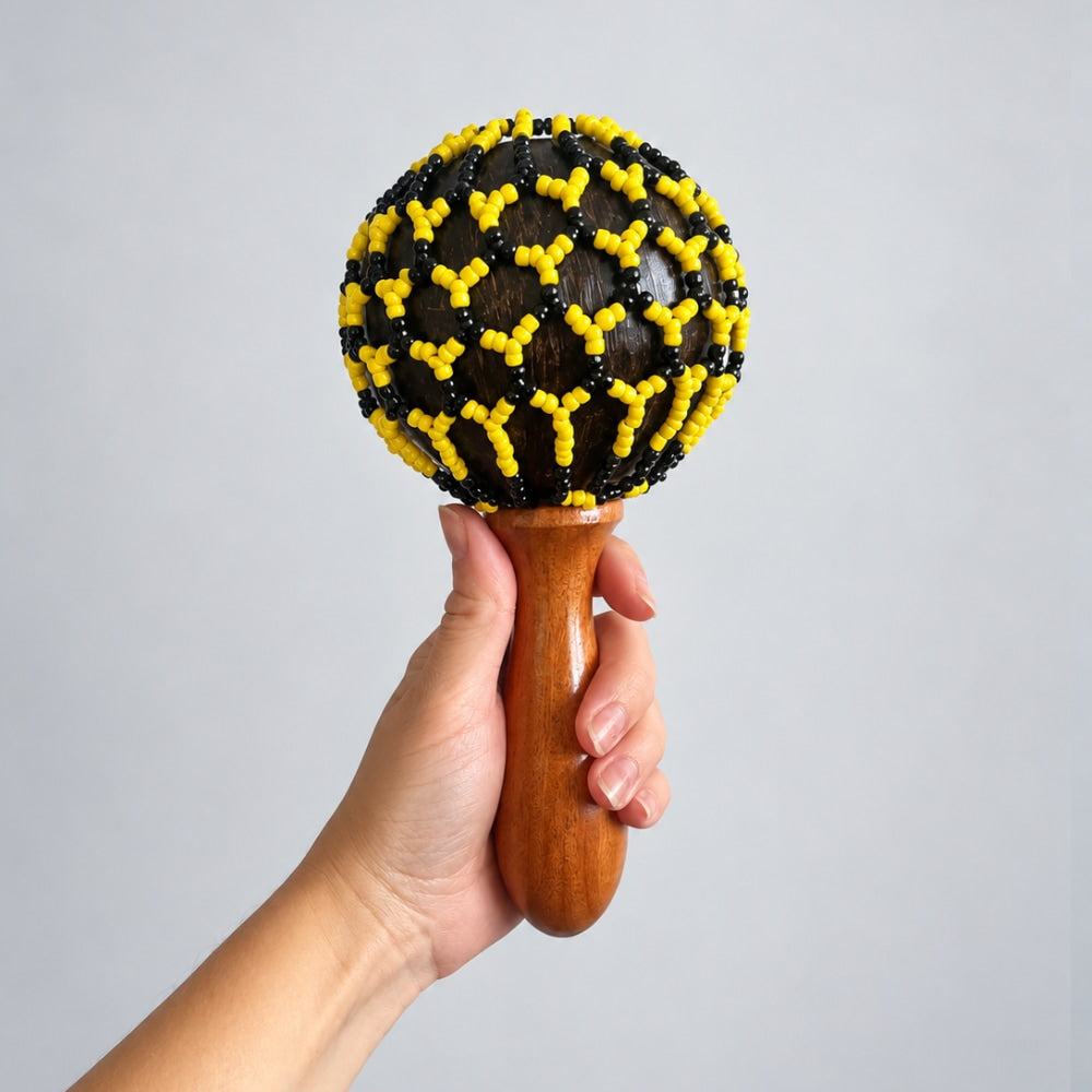 A hand holding a wooden maraca decorated with black and yellow beads, set against a plain light gray background.